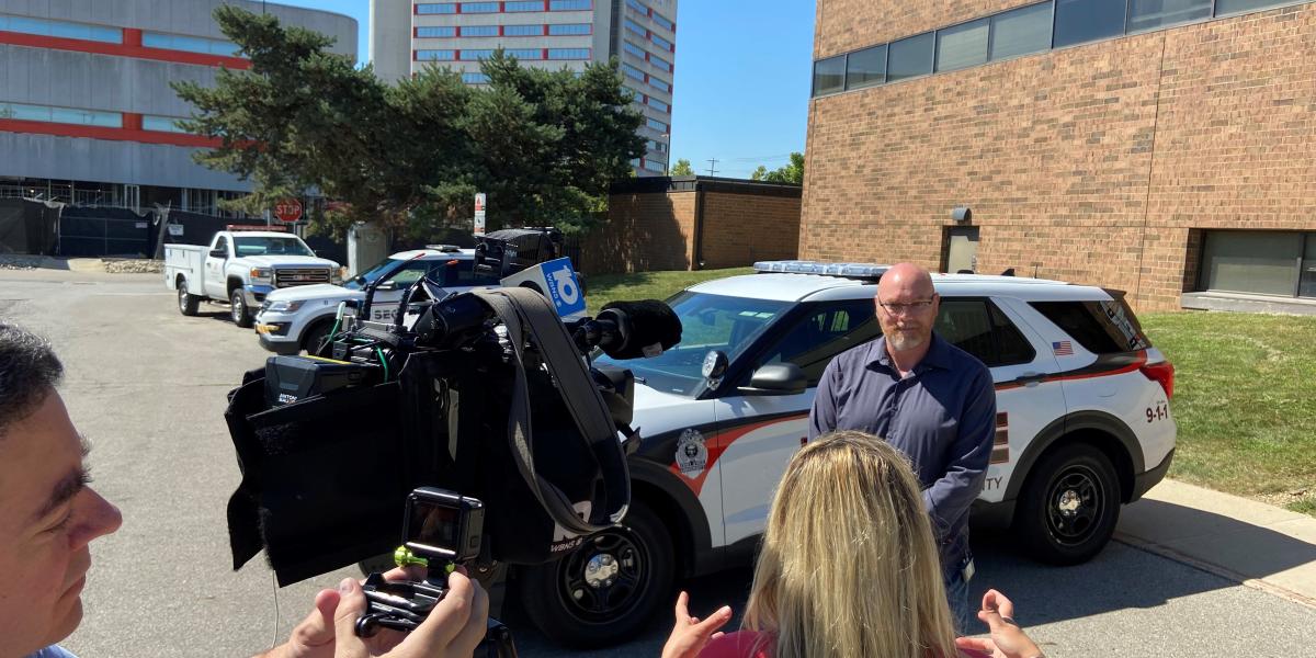 reporter interviews man on camera in front of police car